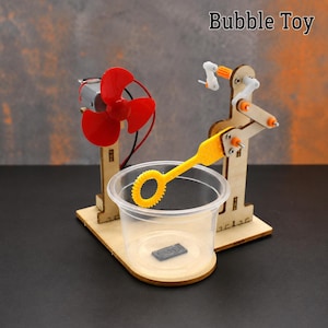 Könnte beinhalten: Ein DIY-Seifenblasen-Spielzeugset mit Holzsockel, kleinem Elektromotor mit rotem Dreiblatt-Propeller und einem mechanischen Arm, der einen gelben Seifenblasenstab über einen transparenten Kunststoffbecher hält. Der Text Bubble Toy ist zu sehen.