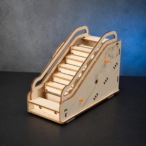 May include: A laser-cut wooden model of a staircase featuring light-colored plywood panels, curved side railings, and small orange plastic accents. The side panels display intricate engraved line patterns and geometric details throughout the structure.