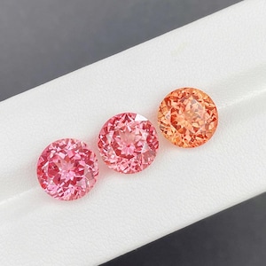 Puede incluir: Se muestran tres gemas redondas en tonos rosa y naranja. Las gemas están facetadas, reflejando la luz y mostrando su estructura interna. Las gemas están dispuestas sobre una superficie blanca, con un fondo gris oscuro.