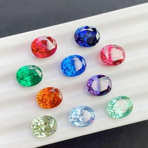 May include: An assortment of oval-cut gemstones in various colors, including red, blue, green, orange, purple, and light blue. The gems are arranged on a white surface, showcasing their facets and brilliance. The gemstones are a mix of different sizes and shades.