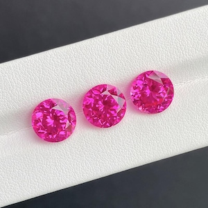 May include: Three round, faceted, hot pink gemstones are displayed on a white surface. The gems have a brilliant cut, reflecting light. The background is a contrasting dark gray and white.