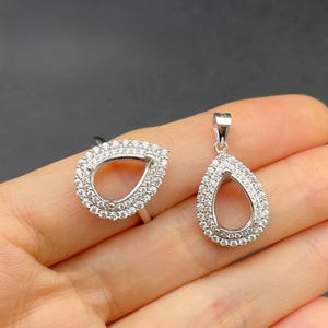 Peut inclure: Un ensemble bague et pendentif en argent. Les deux pièces présentent une forme de goutte d'eau, bordée de petites pierres précieuses transparentes. La bague a un anneau, tandis que le pendentif a une boucle pour une chaîne. Les bijoux sont présentés sur fond neutre.