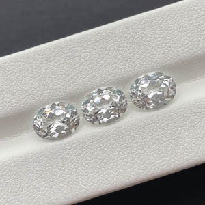 May include: Three oval-cut, clear gemstones with brilliant facets are displayed on a white surface. The gems are arranged in a row, each reflecting light. The stones appear to be diamonds, showcasing a classic jewelry design.