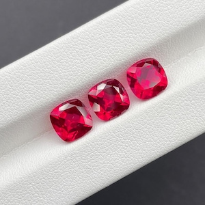 May include: Three faceted, cushion-cut gemstones in a rich red hue. The gems are displayed on a white surface, showcasing their square shape and light reflection. The gems are a deep red colour.