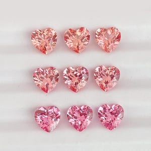 May include: Nine heart-shaped gemstones in varying shades of pink and peach are arranged in a grid pattern. The faceted stones reflect light, showcasing their brilliant cuts. The gems are set against a white background.