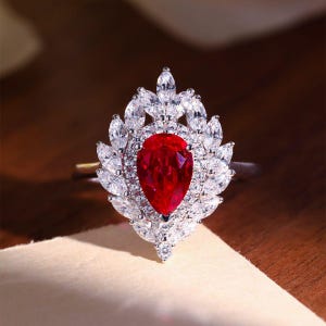 May include: A silver ring featuring a large, pear-shaped, vibrant red gemstone at the center, surrounded by a halo of smaller, clear, marquise-cut stones. The ring's design is ornate and elegant, with a floral-like arrangement of the surrounding stones.