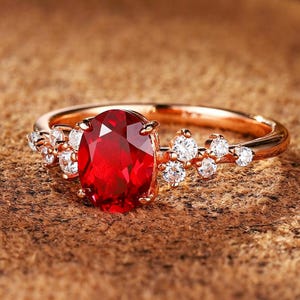Könnte beinhalten: Ein Ring aus Roségold mit einem großen, oval geschliffenen, tiefroten Edelstein in der Mitte. Die Ringschiene ist auf beiden Seiten des zentralen roten Steins mit einer Gruppe kleiner, runder, klarer weißer Steine besetzt, was ein symmetrisches Design ergibt.
