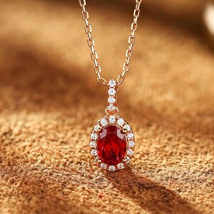 Könnte beinhalten: Eine Halskette aus Roségold mit einem oval geschliffenen, tiefroten Edelsteinanhänger, der von einem Kranz aus kleinen, klaren Steinen umgeben ist. Der Anhänger hängt an einer zarten Roségoldkette mit rechteckigen Gliedern und besetzter Öse.