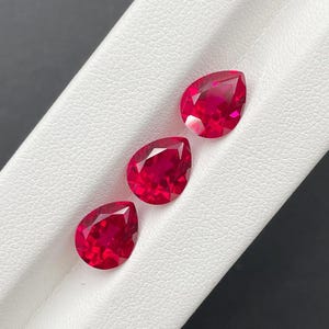 Könnte beinhalten: Drei tropfenförmige, facettierte Rubine. Die Edelsteine haben eine leuchtend rote Farbe und werden auf einer weißen Oberfläche präsentiert. Die Rubine sind diagonal angeordnet und zeigen ihren Schliff und ihre Brillanz. Der Hintergrund ist dunkelgrau.