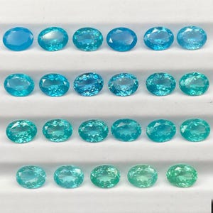 Peut inclure: Une sélection de pierres précieuses de forme ovale, dans des tons de bleu et de vert. Les gemmes sont disposées en rangées, mettant en valeur leur clarté et leurs variations de couleur. Les pierres sont présentées sur une surface blanche.