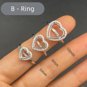 May include: Three silver heart-shaped rings with clear gemstones. The rings are displayed on a finger, with sizes 6mm, 8mm, and 10mm labelled. The text "B - Ring" is in a grey oval in the upper left corner.