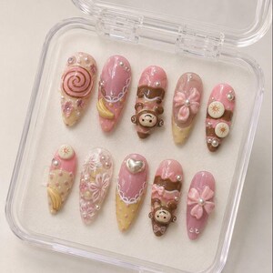 Puede incluir: Un juego de diez uñas artificiales en una caja de plástico transparente. Las uñas presentan varios diseños, incluyendo espirales, flores, un mono y un corazón. Los colores incluyen rosa, marrón y amarillo, con acentos de perlas. Las uñas son en forma de almendra.
