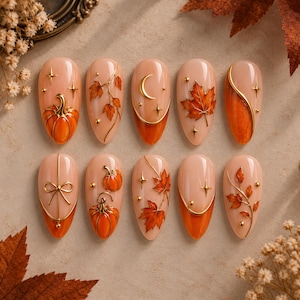 May include: A set of ten almond-shaped press-on nails featuring a nude base with autumn-themed designs. The nails are decorated with raised gold metallic accents, orange pumpkins, maple leaves, crescent moons, stars, and delicate bows.