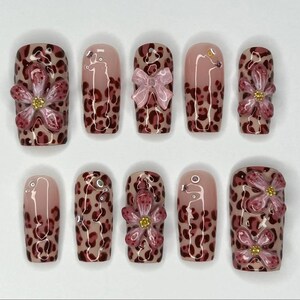 Peut inclure: Un ensemble de dix faux ongles. Les ongles présentent un motif léopard dans des tons de marron et de rose. Certains ongles ont des embellissements de fleurs et de nœuds 3D, tandis que d'autres ont de petits strass argentés. Les ongles sont de forme rectangulaire.