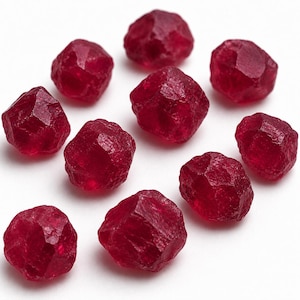 May include: A collection of faceted, deep red gemstones, possibly garnets, arranged on a white surface. Each stone has a rough, irregular shape with multiple facets, catching the light and displaying a rich, saturated color. The gemstones are scattered, creating a visually appealing display.