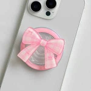 Könnte beinhalten: Ein hellrosa Handygriff mit einer auffälligen Schleife in einem weiß-rosa Vichy-Karomuster. Der runde Griff ist auf der Rückseite eines weißen Smartphones befestigt, bei dem das Kameramodul mit drei Linsen im oberen Bereich zu sehen ist.