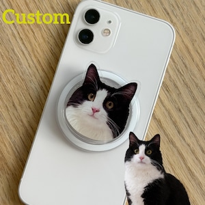 May include: White smartphone with a black and white cat pop socket attached. The pop socket features a close-up image of a cat's face. The word "Custom" is written in yellow on the top left of the phone.