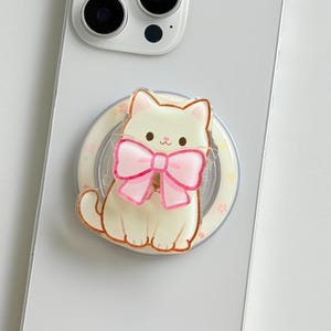 Peut inclure: Un smartphone blanc avec un support de téléphone circulaire fixé au dos. Le support présente une illustration de chat crème avec un grand nœud rose autour du cou, entouré d'un matériau acrylique transparent sur une base circulaire claire.