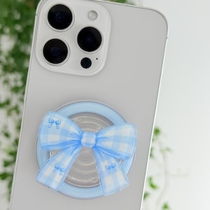 May include: A light blue circular phone grip attached to the back of a smartphone. The grip features a prominent, three-dimensional bow design with a light blue and white gingham check pattern, centered over the circular base of the accessory.