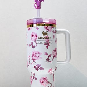 May include: White Stanley tumbler with a floral rose pattern and a pink lid. The tumbler has a white handle and a purple bow on the straw. The text "Stanley" and "LoveShackFancy" are visible.