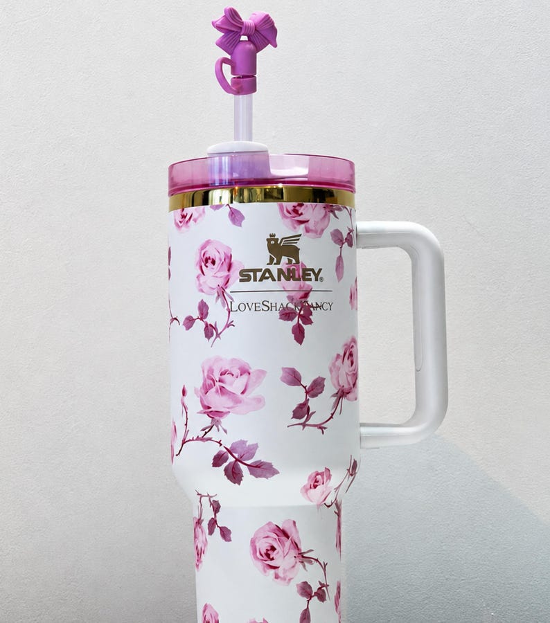 May include: White Stanley tumbler with a floral rose pattern and a pink lid. The tumbler has a white handle and a purple bow on the straw. The text "Stanley" and "LoveShackFancy" are visible.