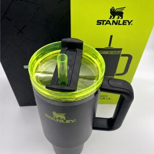 Peut inclure: Un mug de voyage Stanley gris avec une poign&eacute;e noire et un couvercle transparent avec un bord vert fluo. Le couvercle a un curseur noir et une paille verte. Le logo Stanley est imprim&eacute; en vert sur le mug et la bo&icirc;te derri&egrave;re.