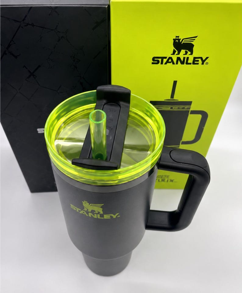 Peut inclure: Un mug de voyage Stanley gris avec une poign&eacute;e noire et un couvercle transparent avec un bord vert fluo. Le couvercle a un curseur noir et une paille verte. Le logo Stanley est imprim&eacute; en vert sur le mug et la bo&icirc;te derri&egrave;re.