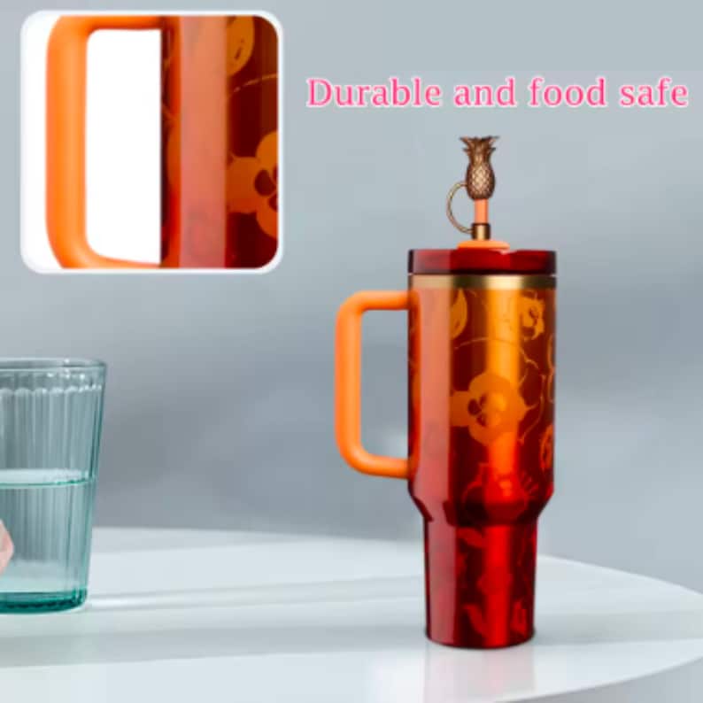 May include: A red and orange insulated travel mug with a handle and a pineapple-shaped lid. The mug features a floral pattern and the text "Durable and food safe". The handle and lid are orange.