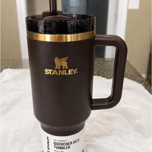 以下が含まれることがあります： ハンドルとストローが付いた、濃い茶色のスタンレークエンチャーH2.0タンブラー。タンブラーには金のバンドとスタンレーのロゴが付いています。ラベルには「THE FLOWSTATE QUENCHER H2.0 TUMBLER 40 OZ | 1.18 L All Day Hydration」と書かれています。