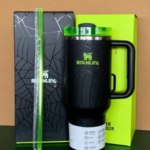 Peut inclure: Gobelet Stanley noir avec couvercle et poign&eacute;e verts, motif toile d'araign&eacute;e. Comprend une paille verte et deux bo&icirc;tes, une noire avec un motif toile d'araign&eacute;e et l'autre vert fluo. Le logo Stanley est visible.