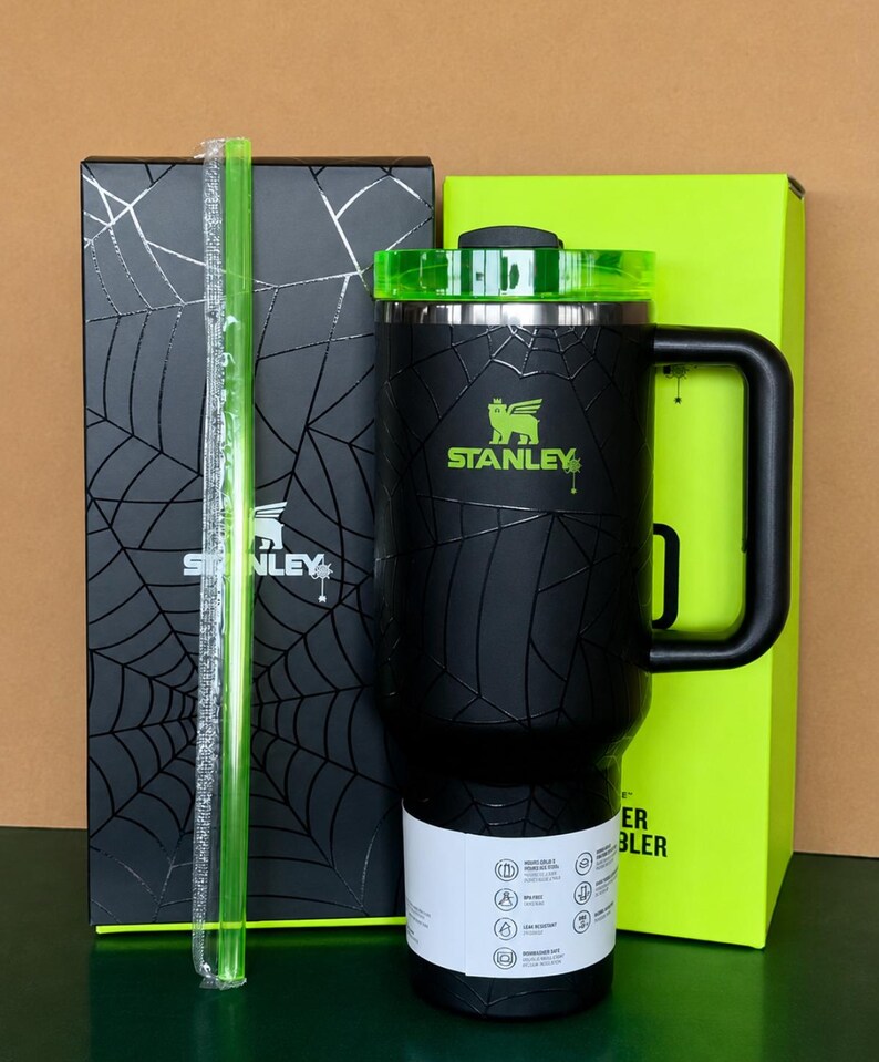 Peut inclure: Gobelet Stanley noir avec couvercle et poign&eacute;e verts, motif toile d'araign&eacute;e. Comprend une paille verte et deux bo&icirc;tes, une noire avec un motif toile d'araign&eacute;e et l'autre vert fluo. Le logo Stanley est visible.