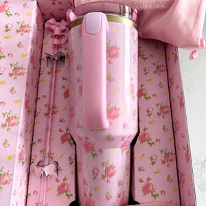 May include: A pink floral-patterned Stanley tumbler with a matching handle and lid, presented in a decorative box. The box and accessories, including a straw and a carrying bag, are also pink. The design features a ribbon with the Stanley and Loveshackfancy logos.