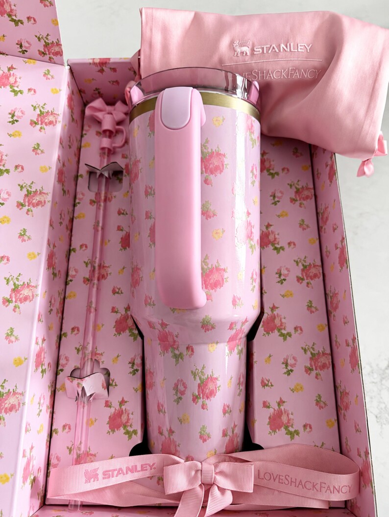 May include: A pink floral-patterned Stanley tumbler with a matching handle and lid, presented in a decorative box. The box and accessories, including a straw and a carrying bag, are also pink. The design features a ribbon with the Stanley and Loveshackfancy logos.