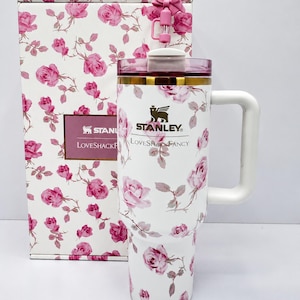 May include: White Stanley LoveshackFancy tumbler with a pink rose pattern, a white handle, and a pink lid. The tumbler is next to a matching gift box. The Stanley logo and LoveshackFancy text are visible.