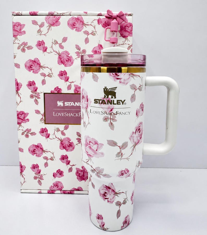 May include: White Stanley LoveshackFancy tumbler with a pink rose pattern, a white handle, and a pink lid. The tumbler is next to a matching gift box. The Stanley logo and LoveshackFancy text are visible.