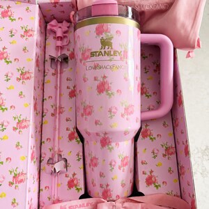 May include: A pink Stanley tumbler with a floral pattern, a matching handle, and a straw, presented in a floral-printed box. The tumbler has the Stanley and LoveShackFancy logos. A pink bow and a pink fabric bag are also included.