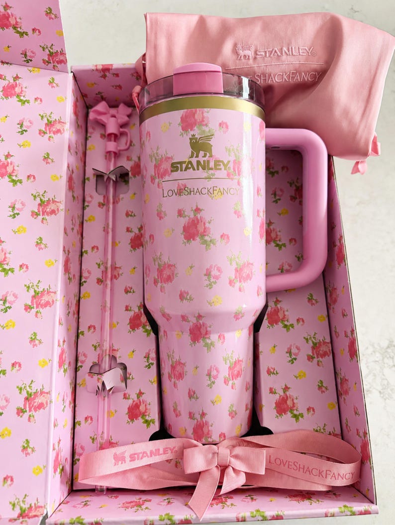 May include: A pink Stanley tumbler with a floral pattern, a matching handle, and a straw, presented in a floral-printed box. The tumbler has the Stanley and LoveShackFancy logos. A pink bow and a pink fabric bag are also included.