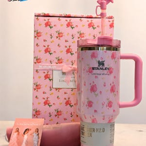 May include: A pink Stanley Quencher H2.0 tumbler with a floral pattern, a matching pink handle, and a pink bow on the straw. The tumbler is part of a gift set that includes a pink box and a card with women in dresses.