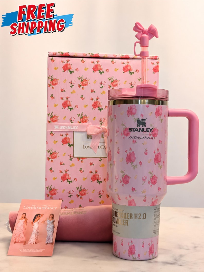 May include: A pink Stanley Quencher H2.0 tumbler with a floral pattern, a matching pink handle, and a pink bow on the straw. The tumbler is part of a gift set that includes a pink box and a card with women in dresses.