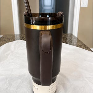 以下が含まれることがあります： ゴールドのバンドとハンドルが付いた濃い茶色のトラベルマグ。マグには透明な蓋と濃い茶色のストローが付いています。マグの底にはテキストが書かれた白いラベルが付いています。マグは飲み物を入れるように設計されています。