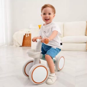 May include: A child's balance bike in a neutral color scheme with orange accents. The bike has a small basket and large wheels. The child is wearing a white t-shirt and blue shorts. The child is smiling.