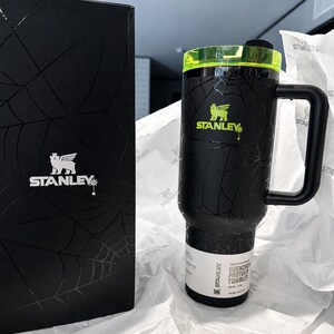 Peut inclure: Gobelet Stanley Quencher Flowstate noir avec un motif toile d'araign&eacute;e, un couvercle vert n&eacute;on et une poign&eacute;e noire. Le gobelet porte le logo Stanley en vert n&eacute;on. Une bo&icirc;te noire avec le logo Stanley et un motif toile d'araign&eacute;e est &agrave; c&ocirc;t&eacute; du gobelet.