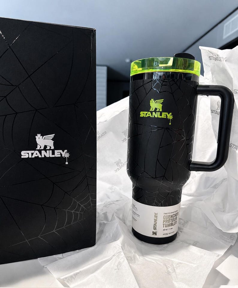 Peut inclure: Gobelet Stanley Quencher Flowstate noir avec un motif toile d'araign&eacute;e, un couvercle vert n&eacute;on et une poign&eacute;e noire. Le gobelet porte le logo Stanley en vert n&eacute;on. Une bo&icirc;te noire avec le logo Stanley et un motif toile d'araign&eacute;e est &agrave; c&ocirc;t&eacute; du gobelet.