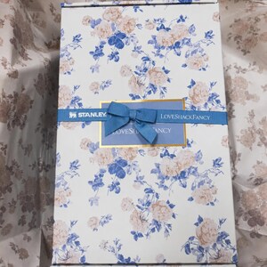 May include: A rectangular box with a floral pattern of blue and peach roses on a white background. A blue ribbon with a bow and the text "LOVESHACKFANCY" and "STANLEY" is wrapped around the box.