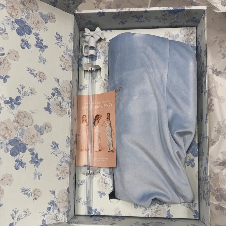 May include: An open box with a floral pattern in white and blue, containing a light blue garment, a long glass item with a bow, and a brochure. The brochure has the text "LAUNCH PARTY" and images of women in dresses.