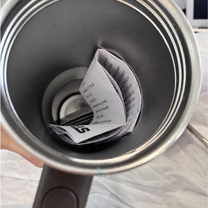 以下が含まれることがあります： ダークブラウンのハンドルが付いたステンレス製のトラベルマグ。マグの内側には、テキストが書かれた折り畳まれた白い紙が見えます。マグの内側は濃いグレー色です。