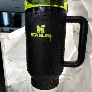 Peut inclure: Gobelet Stanley Quencher Protour noir avec un motif en toile d'araign&eacute;e textur&eacute;. Le gobelet a une poign&eacute;e noire et un couvercle vert fluo. Le logo Stanley est en vert fluo.