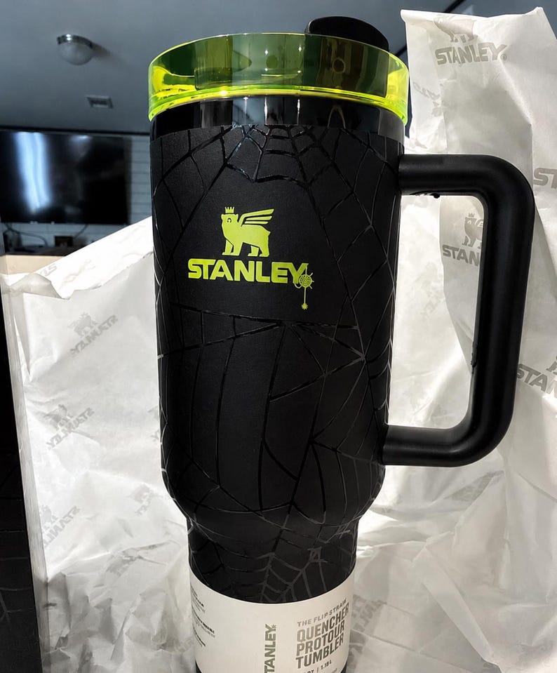 Peut inclure: Gobelet Stanley Quencher Protour noir avec un motif en toile d'araign&eacute;e textur&eacute;. Le gobelet a une poign&eacute;e noire et un couvercle vert fluo. Le logo Stanley est en vert fluo.
