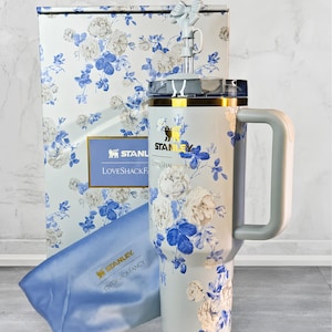 May include: A Stanley Loveshackfancy tumbler with a handle, straw, and lid, featuring a floral design in white and blue. The tumbler is accompanied by a matching gift box and a blue cloth. The Stanley logo is visible.