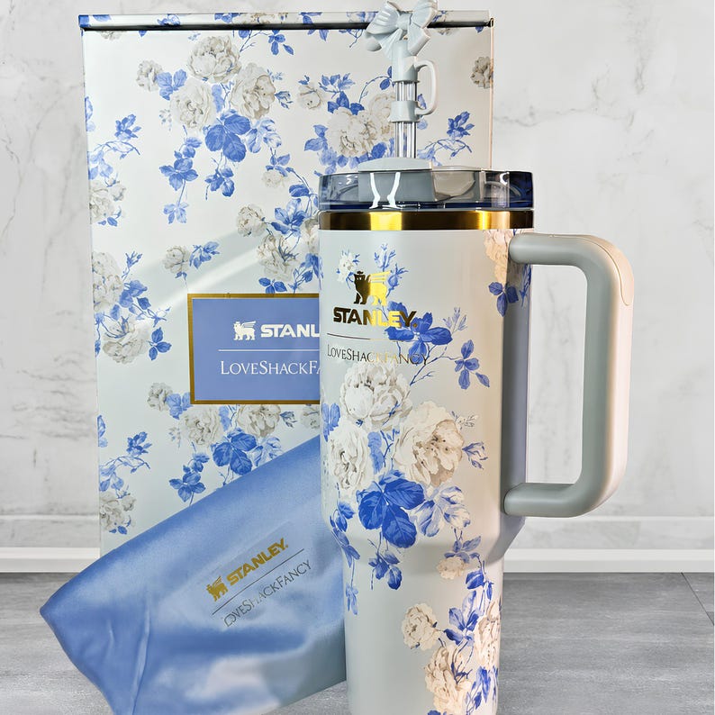 May include: A Stanley Loveshackfancy tumbler with a handle, straw, and lid, featuring a floral design in white and blue. The tumbler is accompanied by a matching gift box and a blue cloth. The Stanley logo is visible.